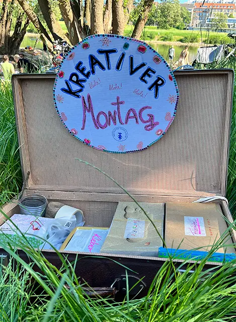 Ein alter Koffer im Gras, darin liegt Bastelmaterial, obenauf ein handgemaltes Schild „Kreativer Montag“