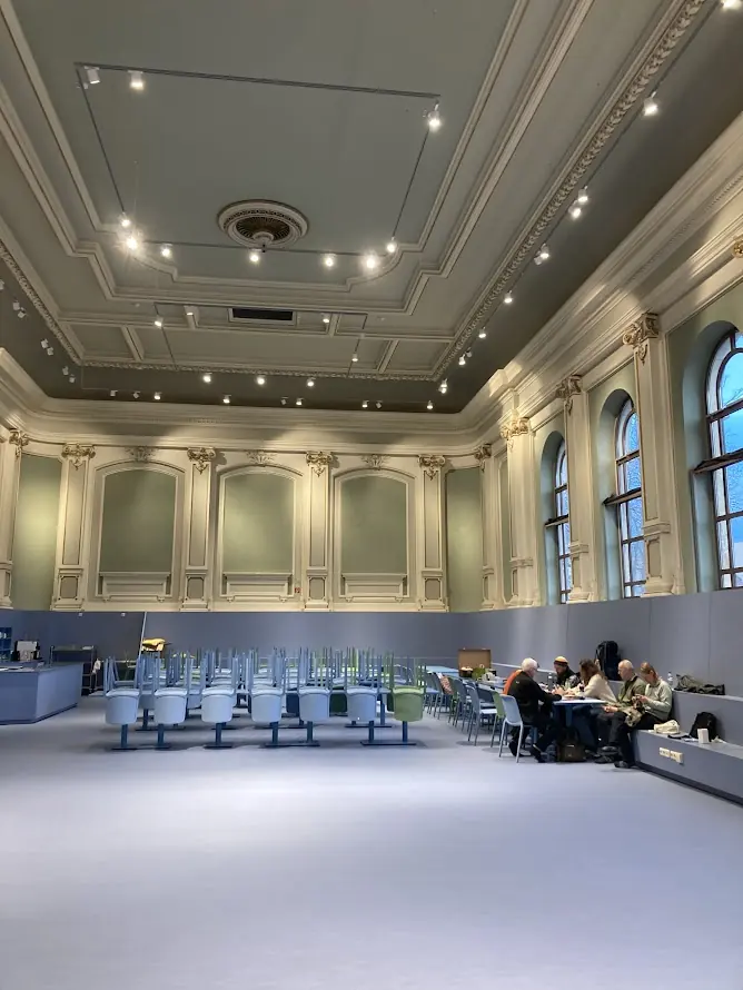 Hoher historischer Saal der Kulturwerkschule mit Stuckdecke und großen Fenstern, an einem Tisch sitzt eine kleine Gruppe
