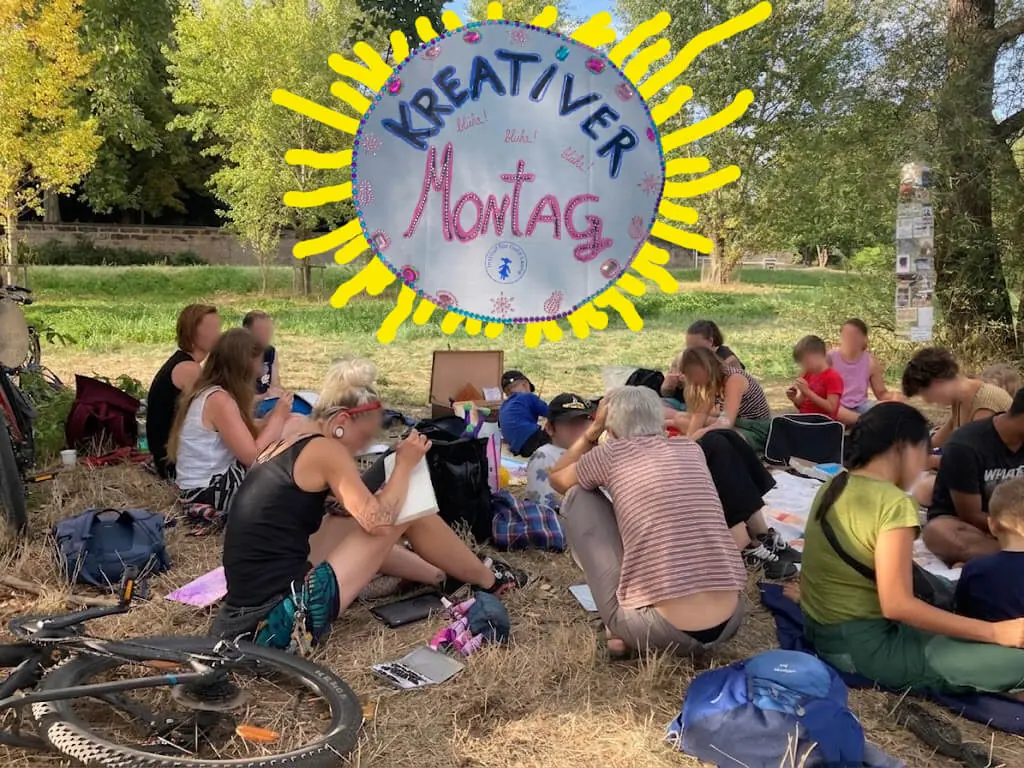 Gruppe sitzt draußen auf Decken an der Prießnitzmündung, darüber ein handgemaltes Schild „Kreativer Montag“ mit Sonnenstrahlen