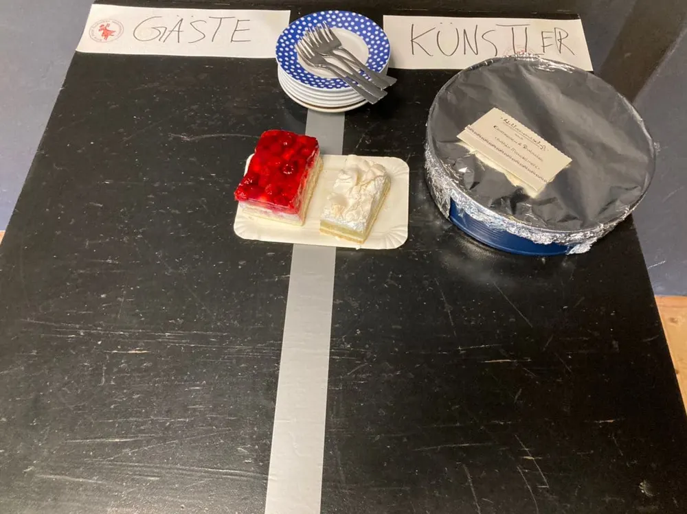 Kuchentisch mit den Schildern „Gäste“ und „Künstler“, dazwischen eine Klebeband-Linie; auf jeder Seite steht ein Stück Kuchen
