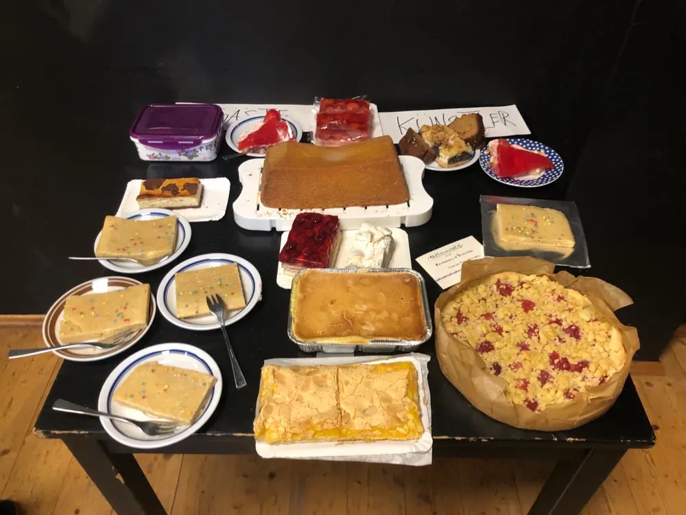 Voller Kuchentisch mit Blechkuchen, Torten und einzelnen Stücken auf Tellern, dahinter die Schilder „Gäste“ und „Künstler“