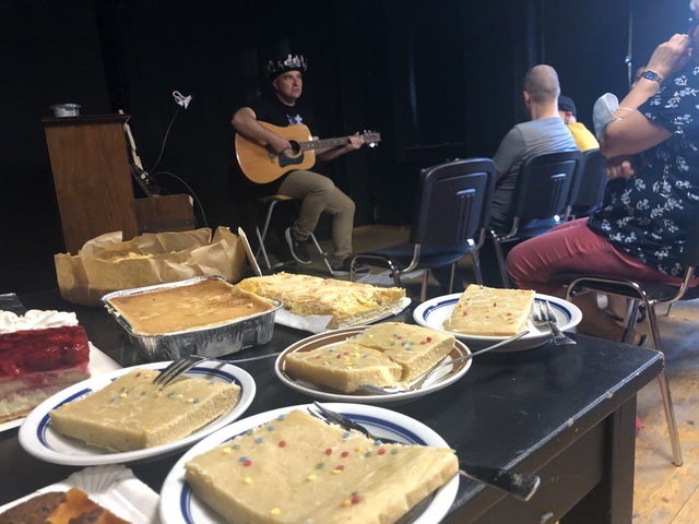 Ein langer Tisch voller selbstgebackener Kuchen, dahinter singt ein Musiker mit Gitarre für das sitzende Publikum.