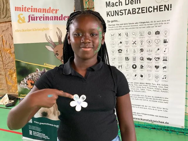 Ein Mädchen zeigt auf das Kunstabzeichen an ihrem Shirt, eine weiße Blüte.