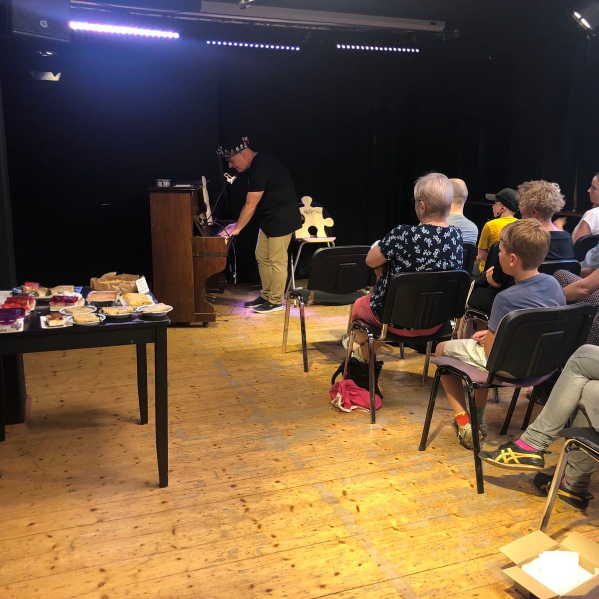 Blick in den Saal: links ein Tisch mit Kuchen, rechts sitzen Kinder und Erwachsene auf Stühlen und schauen zur Bühne